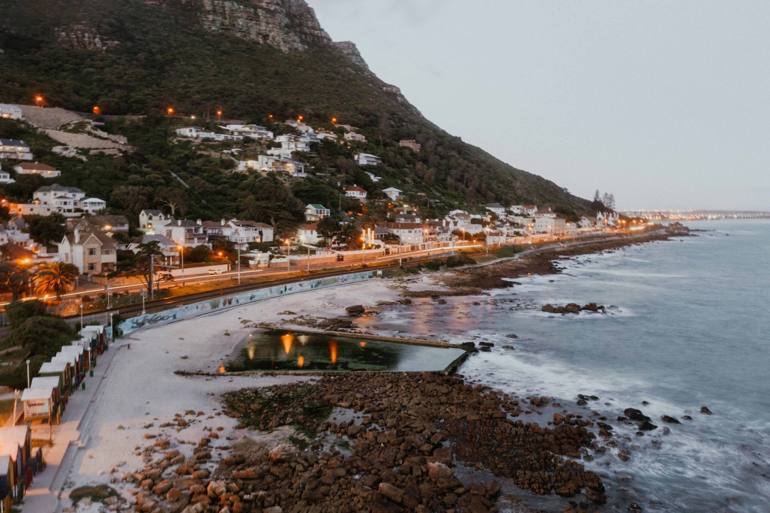 Scenic view of Cape Town coastline with houses on the hillside, ocean waves, and evening lights at sunset